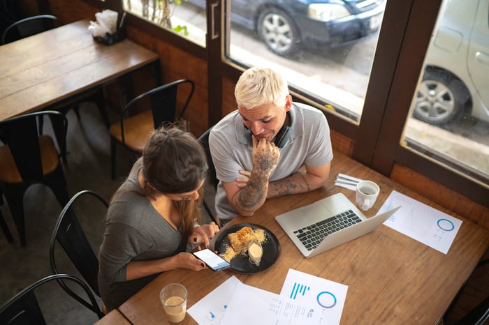 People working at restaurant table with food, paperwork, and a laptop.