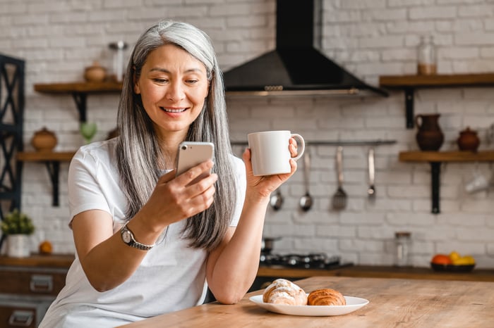 A person holds a coffee mug in a kitchen and smiles at a smartphone. 
