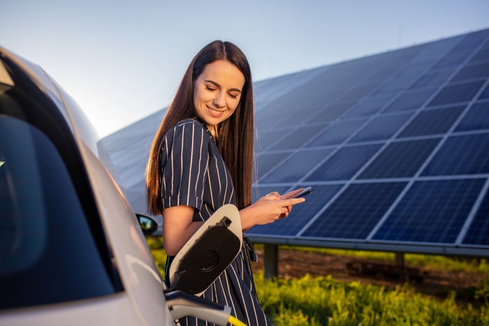 Smiling woman checks her phone while waiting for her electric car to charge.