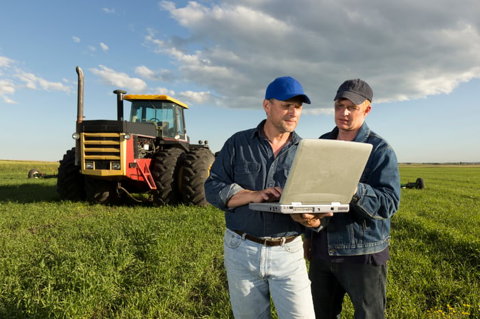 Two people using laptop in the middle of crops with heavy tractor in background