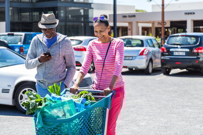 A person pushing a wagon filled with groceries in a parking lot with another person standing next to them.