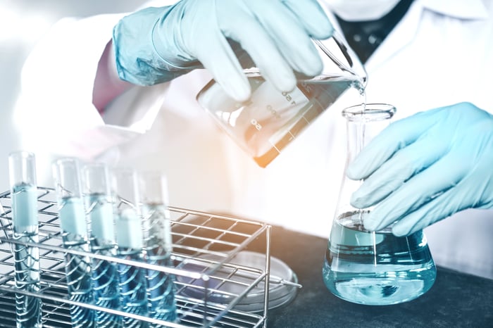 A person in a white lab coat is pouring a chemical solution from a beaker into a flask. 