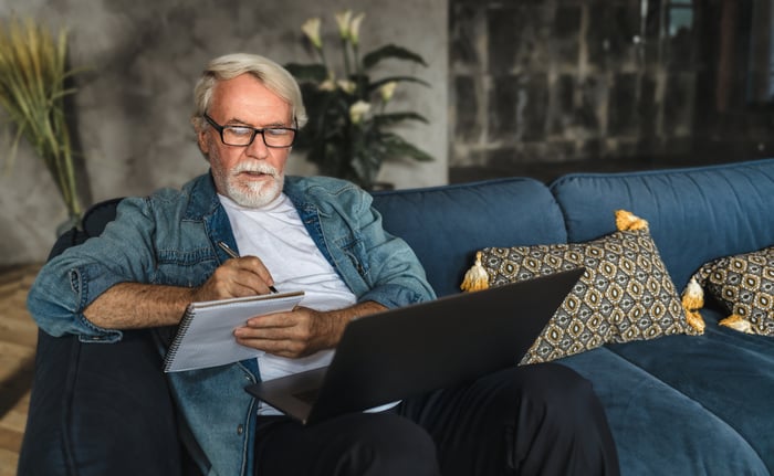 Senior with laptop on lap writing note in notebook.