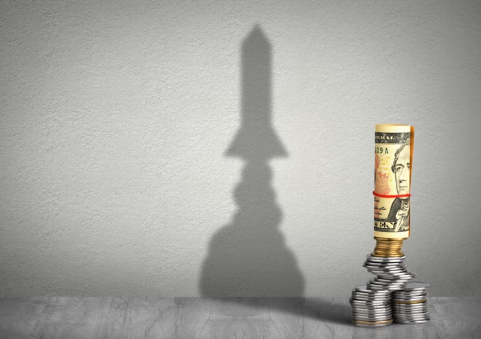 A stack of money casting a shadow that looks like a rocket ship in the background.