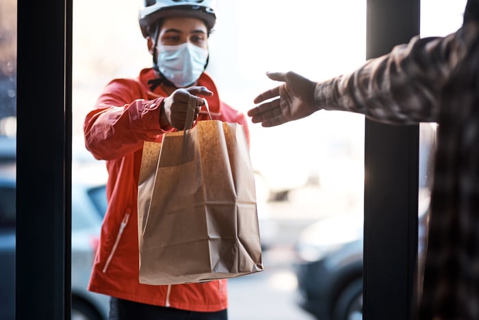 A masked person in a doorway handing over a bag.