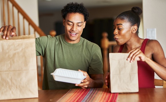 Two people at a table with brown paper bags and a takeout food box.