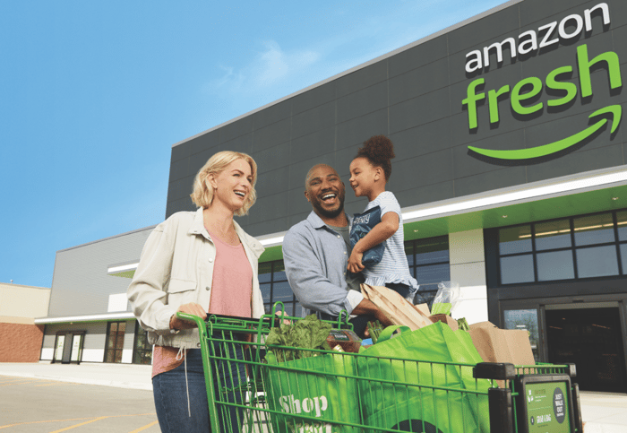 Family smiling while pushing a shopping cart outside an Amazon Fresh store.