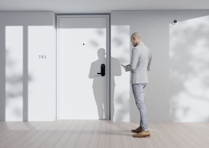 A man approaches an apartment door while looking at his smartphone. The person is using the smartphone to unlock a Latch-equipped door 