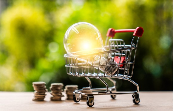A miniature shopping cart with a light bulb inside its basket with stacks of coins in the background.
