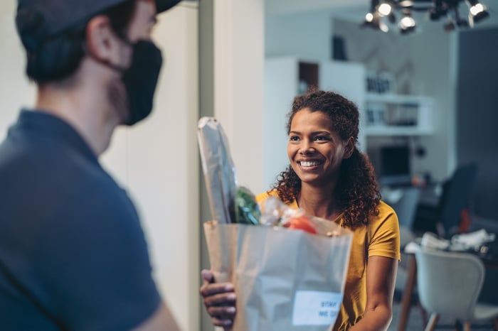 A masked person handing a bag of groceries to another person.