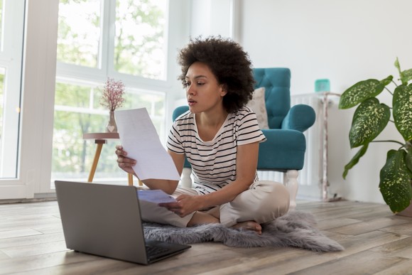 An investor sits on the floor in a living room and works on a laptop.
