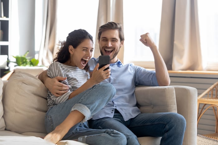 Two people sitting on a couch looking at a phone and cheering.