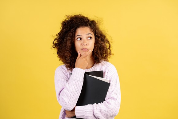 A young person holds notebooks while holding her chin in thought.
