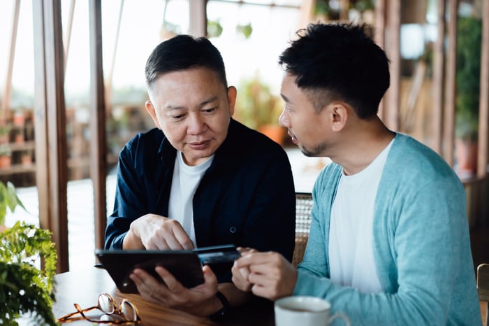 One person explains something to another while seated at a table and pointing to a tablet.