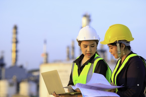 Two engineers talking while looking at papers and a laptop, with an oil refinery in the background.