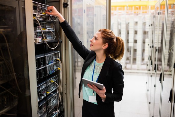 An engineer holding a tablet while checking wires plugged into a data center server tower.