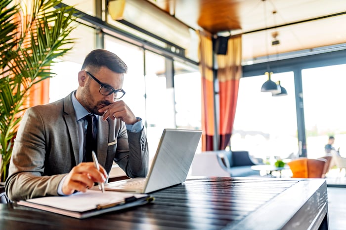 A person in a business suit with a serious expression looking at a laptop and holding a pen.