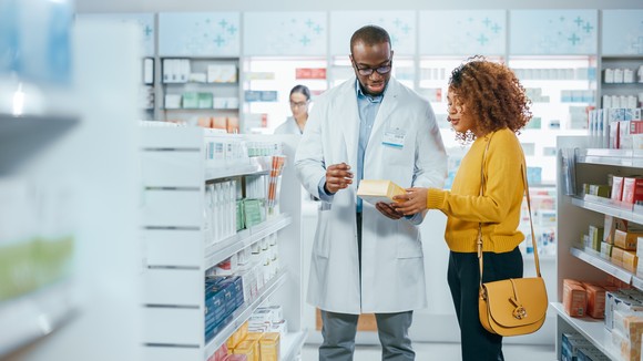 Person in lab coat at pharmacy talking with another person about a product.