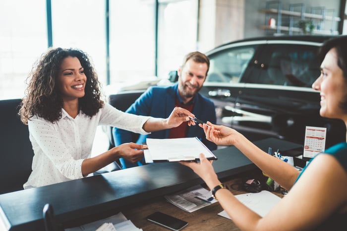 A person hands a clipboard and pen to another person in a car dealership.