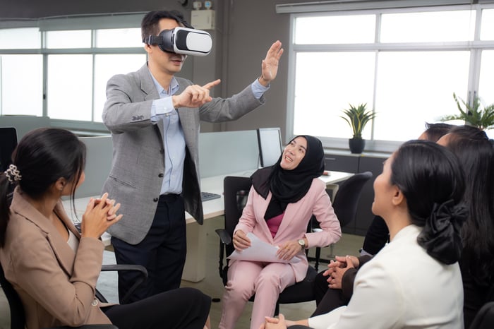 An office worker stands up and wears a VR headset as co-workers observe.