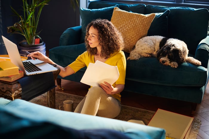 Person sitting on the floor looking at a laptop and documents while dog is resting on a couch in the background.