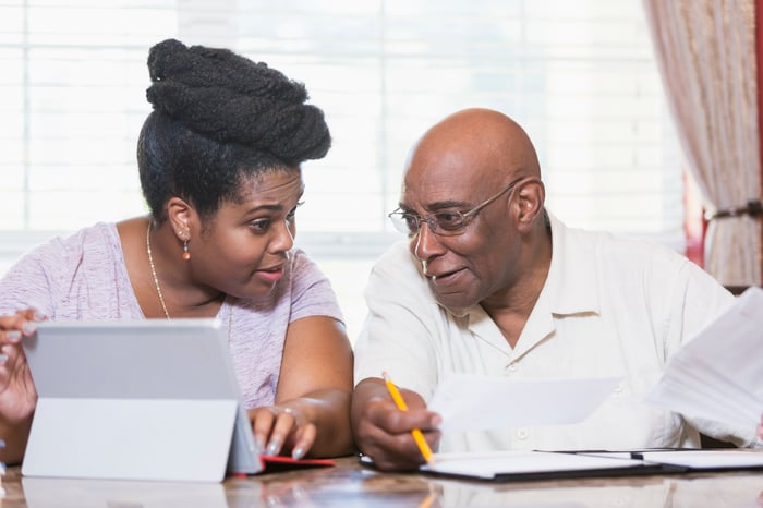 Two people sitting at a table with a tablet, paper, and pencil.