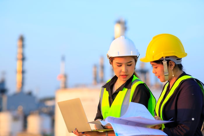 Two engineers having a discussion with an oil refinery in the background.