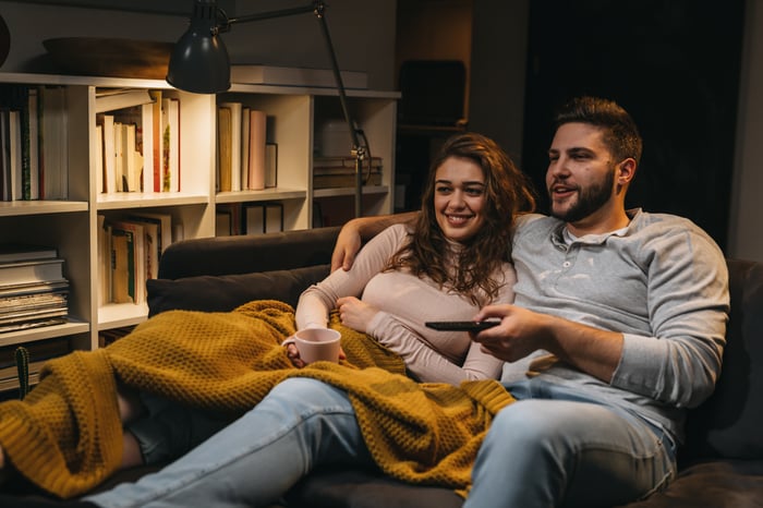 A smiling couple laying on the couch watching a movie, with one of them flicking channels using a remote.