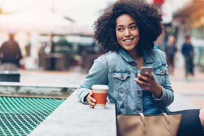 A smiling young person with a smartphone and holding a coffee cup while sitting at a counter.