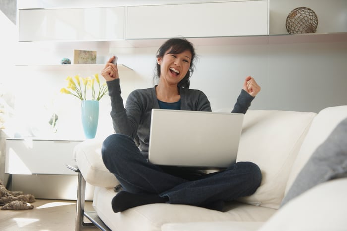 A person holding a credit card and sitting on a couch with a laptop, extremely happy about an online purchase.