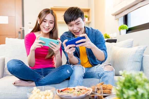 Two young people playing on smartphones while sitting on a couch with plates of food in front of them.