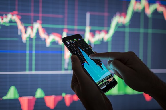 Finger pushes phone showing a stock chart before a larger screen also showing a stock chart.