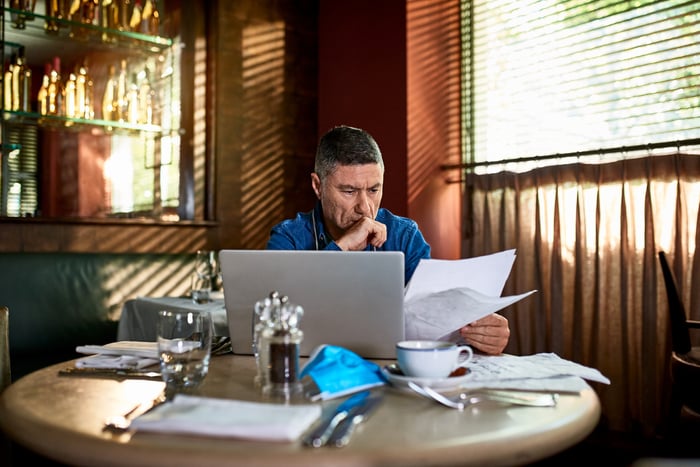 A concerned-looking person seated in a cafe with a laptop while reading from some papers.