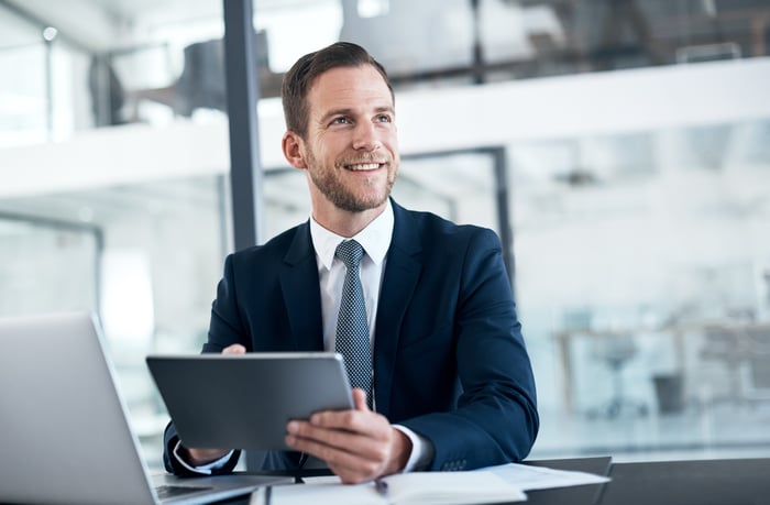 A smiling person in a business suit sitting at a desk and holding a tablet.