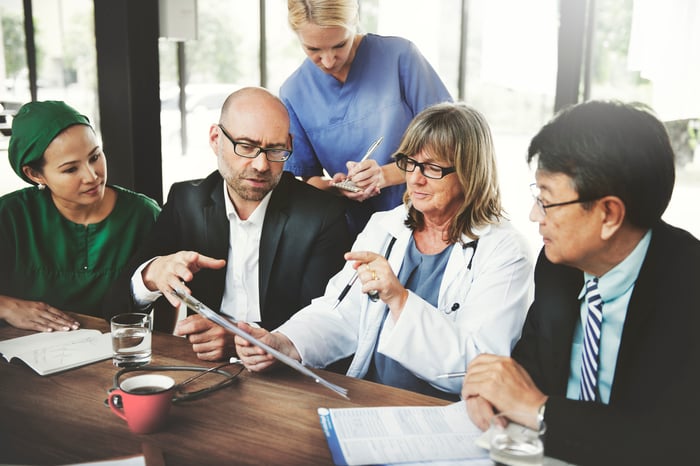 Four people sit at a table reviewing a clipboard, while another person stands behind them taking notes. The person holding the clipboard is wearing a white medical coat and a stethoscope. 