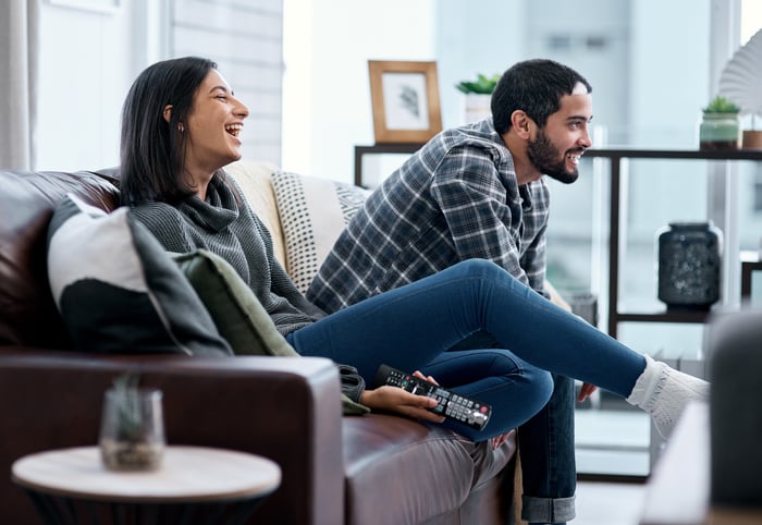 Two people sit on a couch. One person is holding a television remote.