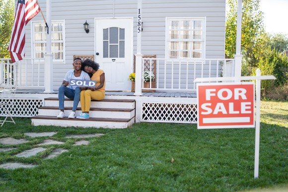 A young couple sits outside their house holding a sign that says "sold."