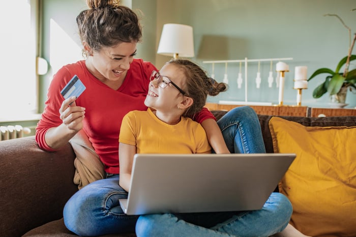 An adult and child smile at each other as they sit on a couch at home and shop online.