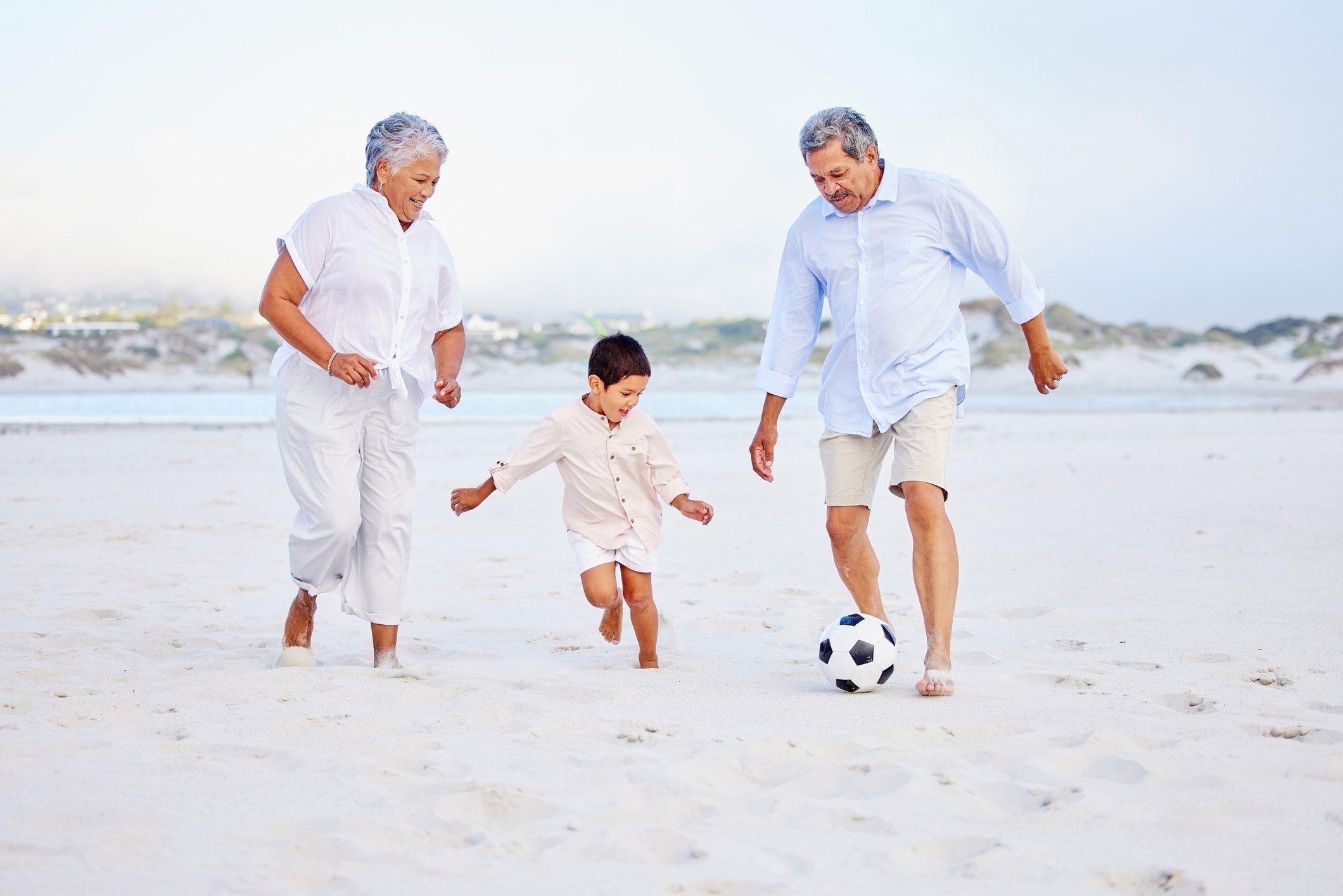 two adults playing soccer with young child on beach