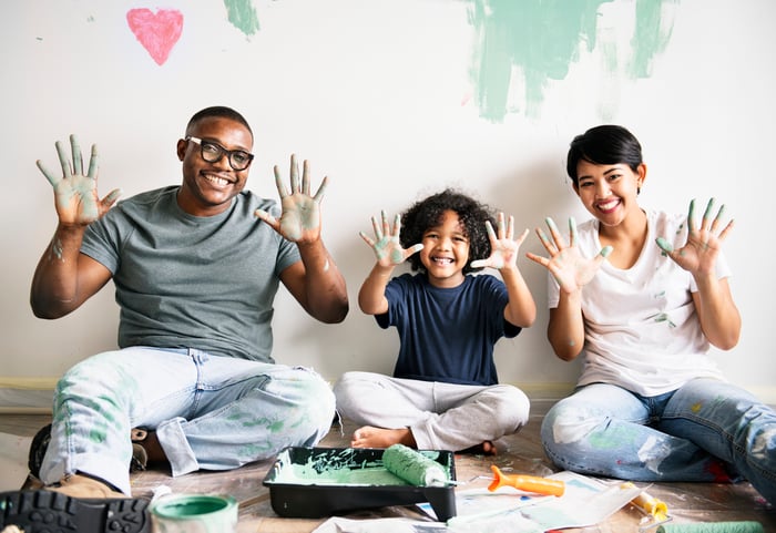 Three people painting a room hold up their hands, which are covered in paint.