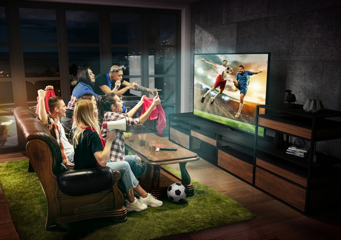 Friends watching a soccer game on TV in a living room on a rug made of grass turf.