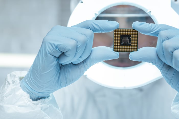 A technician in a full cleanroom suit holds up a square semiconductor chip.