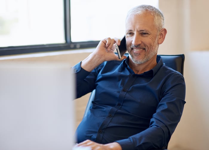 A smiling person at a computer holding a phone.