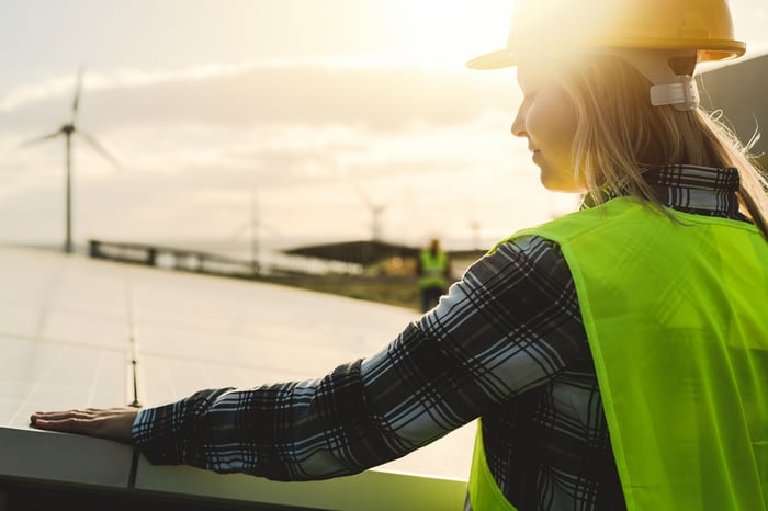 A person installing a solar panel near a wind farm.