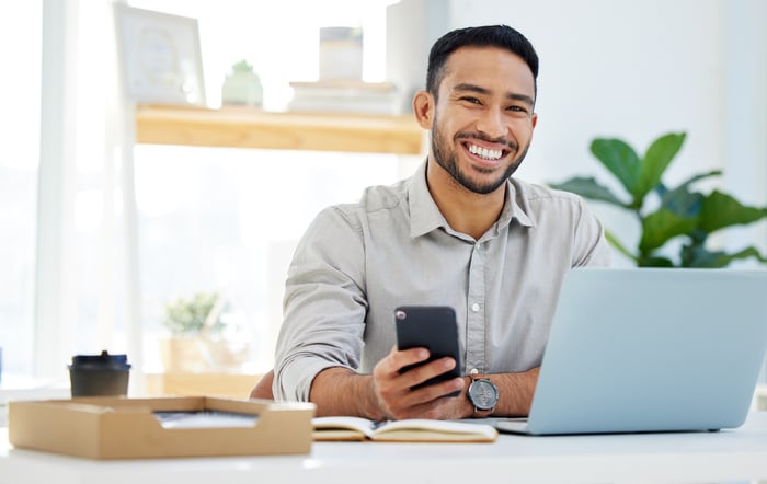 A smiling person at a laptop holding a phone.