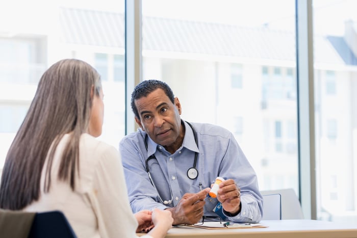 A doctor points to a pill bottle while explaining it to a patient while sitting at a table in a clinic.