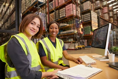 two warhouse workers at computer desk smiling toward camera