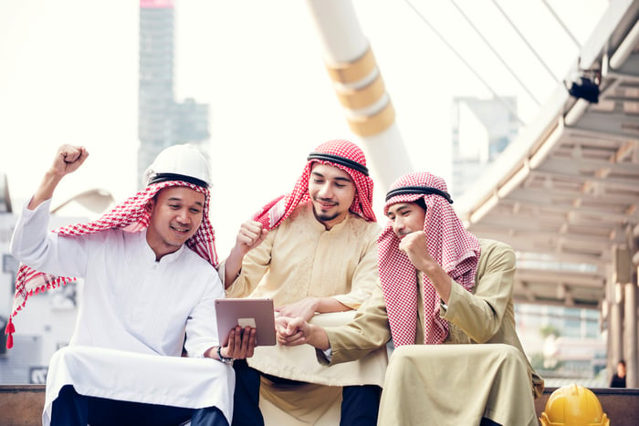 Three Arab men in traditional garb reading news on a tablet and cheering.