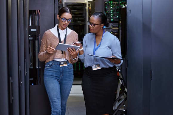 Two techies working on a database on a tablet at a server farm.