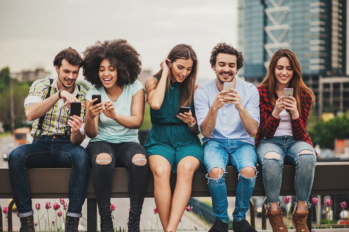 A number of people sitting on a bench and smiling while looking at smartphones.